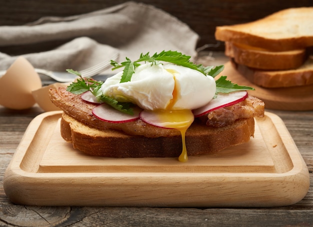 Sandwich sobre una rebanada de pan blanco tostado con huevos escalfados, hojas verdes de rúcula y rábano