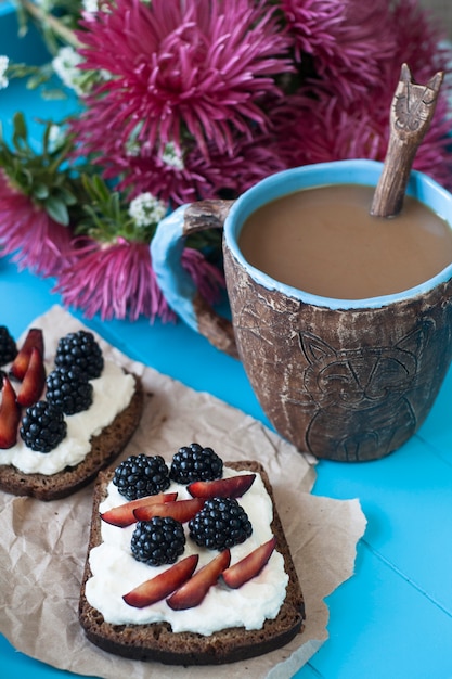 Sandwiche mit süßem Käse und Beeren, einem Tasse Kaffee und einem Blumenstrauß von Astern in der blauen Holzoberfläche