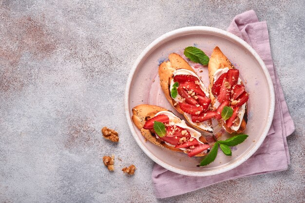 Sándwiches abiertos con fresas, queso suave menta y nuez en placa de cerámica sobre un fondo de piedra gris claro. Verano y comida de dieta saludable, concepto de comida vegetariana. Vista superior.