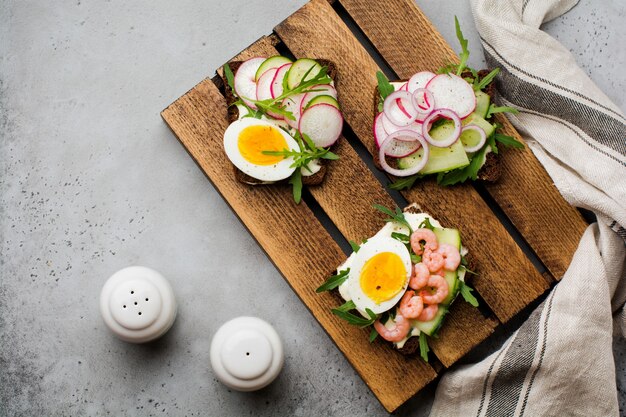 Sándwiches abiertos en pan de centeno oscuro con huevos, camarones, rábanos, pepino, queso crema y rúcula para el desayuno en la vieja superficie oscura