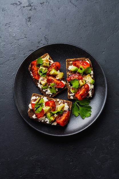 Sándwiches de aguacates, tomates cherry y queso feta con salsa balsámica y cilantro sobre superficie negra oscura. Vista superior.