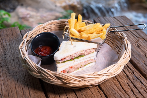 Foto sándwiches de atún servidos con papas fritas y salsa de tomate, dispuestos en una hermosa canasta de ratán, colocados sobre una mesa de madera.