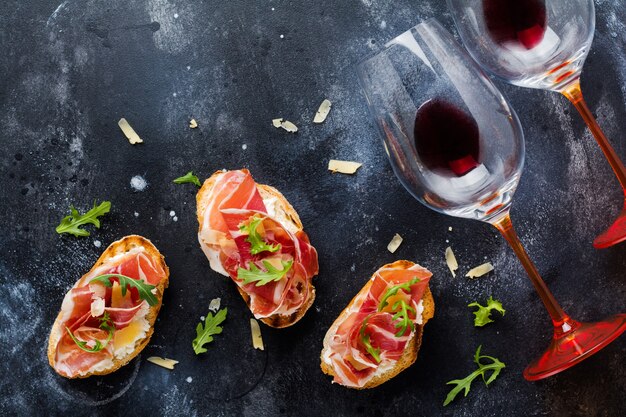 Sándwiches de jamón abierto, rúcula y queso duro, servidos en un soporte de madera con una copa de vino tinto sobre una vieja superficie oscura de hormigón