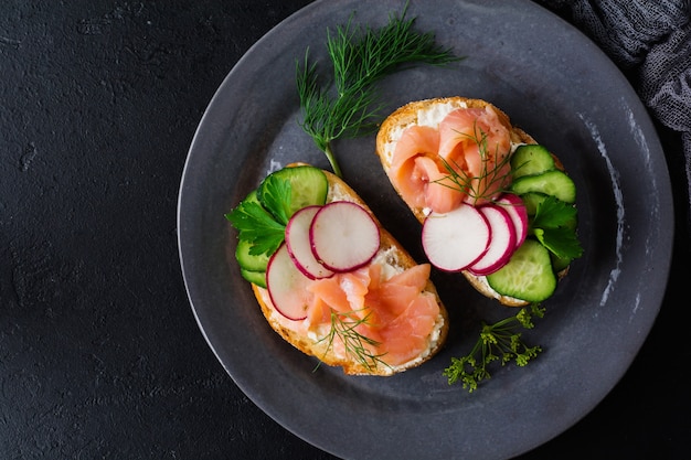 Sandwiches mit geräuchertem rosa Lachs, Radieschen, Gurke und Frischkäse auf grauer Keramikplatte und Textiloberfläche