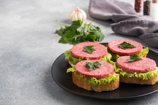 Sandwiches mit Salatblättern und geschnittener Salamiwurst auf schwarzem Teller. Platz kopieren