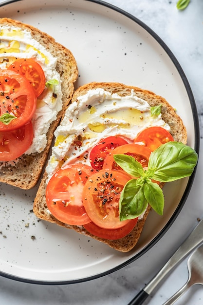 Foto sándwiches o tostadas con tomates, queso crema, aceite de oliva y albahaca sobre un fondo de mármol blanco