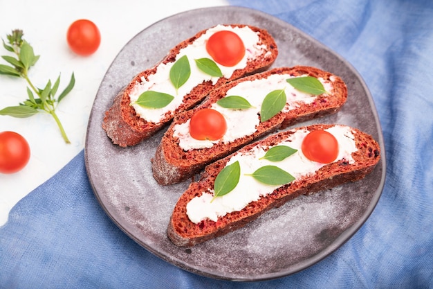 Sándwiches de pan de remolacha roja con queso crema y tomates sobre fondo de hormigón blanco y textil de lino azul. Vista lateral, enfoque selectivo, de cerca.