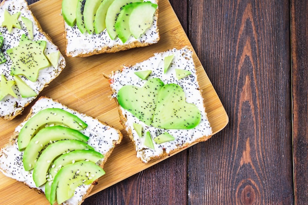 Sándwiches de pan con rodajas, estrellas, corazones de aguacate y requesón sobre fondo de madera.