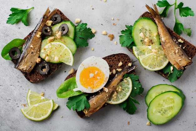 Sándwiches de pescado con espadines, pepino, lima, huevos duros, hojas de perejil y mango en pan de centeno sobre una superficie de hormigón viejo gris. Enfoque selectivo