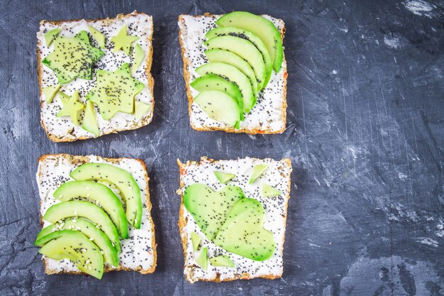 Sándwiches de rebanadas de pan, estrellas, corazones de aguacate y requesón fondo gris oscuro de mármol