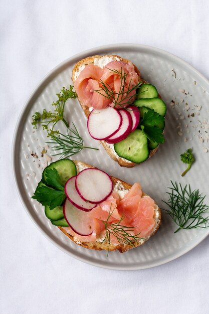 Sándwiches con salmón rosado ahumado, rábano, pepino y queso crema sobre placa de cerámica gris y superficie textil