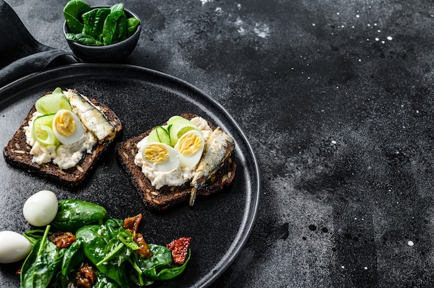 Sandwiches con sardinas, huevo, pepino y queso crema, guarnición de ensalada con espinacas y tomates secos. Fondo negro. Vista superior. Copia espacio