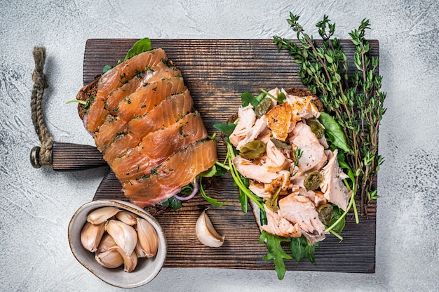 Sandwichtoast mit heißem und kaltem Räucherlachs, Rucola und Frischkäse auf einem Holzbrett. Weißer Hintergrund. Ansicht von oben.