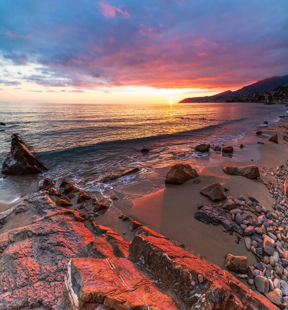 Sanremo, Riviera dei Fiori, Ligurien, Italien. Scenis-Felsen und -Kieselsteine am Strand, die von Sonnenuntergangslicht wunderschön beleuchtet werden. Dramatischer bunter Himmel, der das Meer wellenartig bewegt.