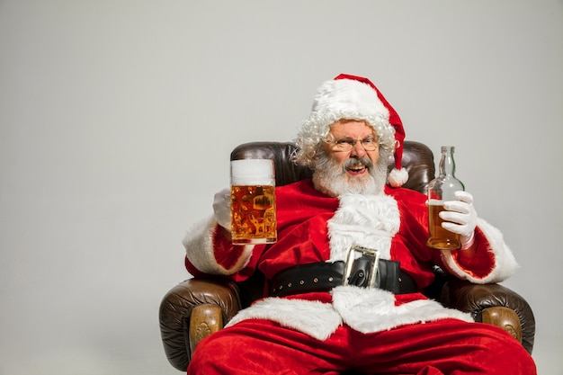 Santa Claus bebiendo cerveza sentado en un sillón, felicitando, parece borracho y feliz. Modelo masculino caucásico en traje típico. Año nuevo 2020, regalos, vacaciones, humor de invierno. Copyspace para su anuncio.