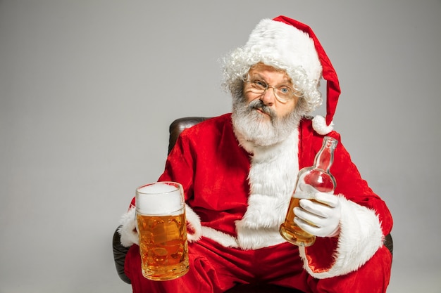 Santa Claus bebiendo cerveza sentado en un sillón, felicitando, parece borracho y feliz. Modelo masculino caucásico en traje típico. Año nuevo 2020, regalos, vacaciones, humor de invierno. Copyspace para su anuncio.