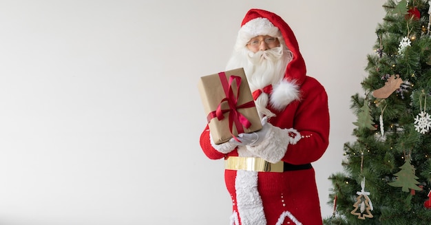 Santa Claus se encuentra cerca del árbol de Navidad sosteniendo una caja con un regalo en sus manos.