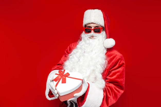 Santa Claus en gafas tiene una caja de regalos en forma de corazón sobre un fondo de color