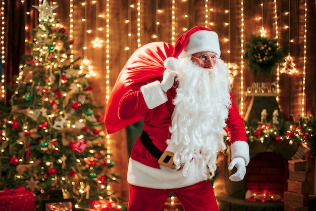 Santa Claus mit einem großen Sack Geschenken an seinem Raum zu Hause nahe Weihnachtsbaum