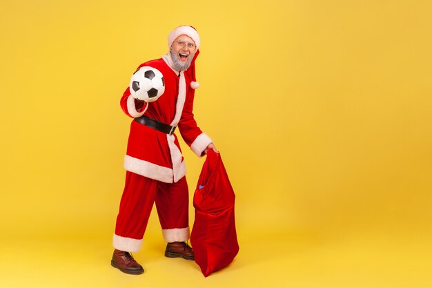 Santa claus de pie con balón de fútbol y gran bolsa roja con regalos de Navidad.