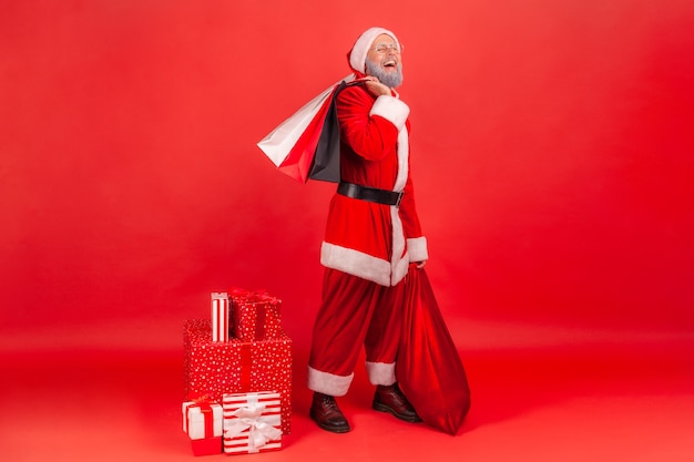 Santa claus de pie con bolsas de regalos y regalos de Navidad, mirando a la cámara.