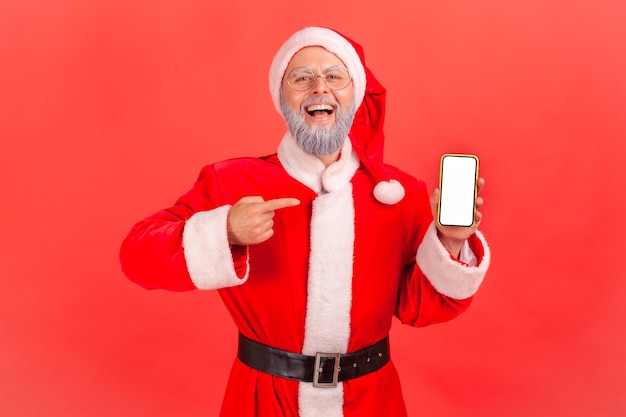 Santa claus de pie con el teléfono inteligente en las manos, apuntando a la pantalla en blanco con el dedo índice.