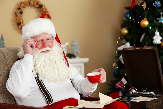 Santa Claus sentado con cartas de niños en una silla cómoda cerca de la chimenea en casa
