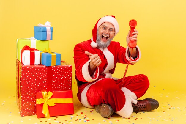 Santa claus sentado regalos de Navidad, mostrando el pulgar hacia arriba y hablando por teléfono fijo.