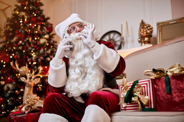 Santa Claus sentado en el sofá y hablando por teléfono móvil cerca de la chimenea y el árbol de Navidad con regalos. Año nuevo y feliz Navidad, felices fiestas concepto