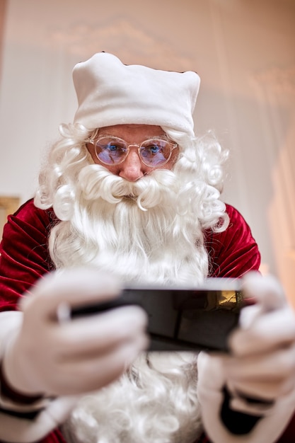 Santa Claus sentado en el sofá y hablando por teléfono móvil cerca de la chimenea y el árbol de Navidad con regalos.