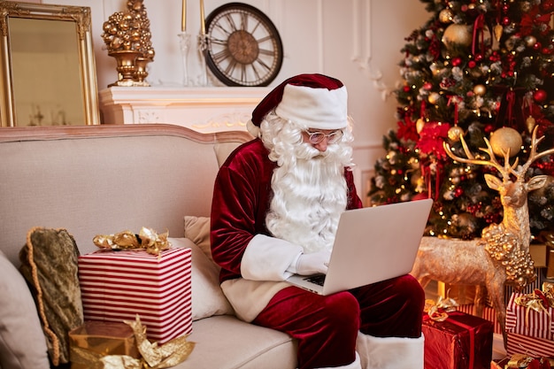 Santa Claus sentado en su casa y leyendo el correo electrónico en la computadora portátil con solicitud de Navidad o lista de deseos cerca de la chimenea y el árbol con regalos. Feliz Navidad, felices fiestas concepto