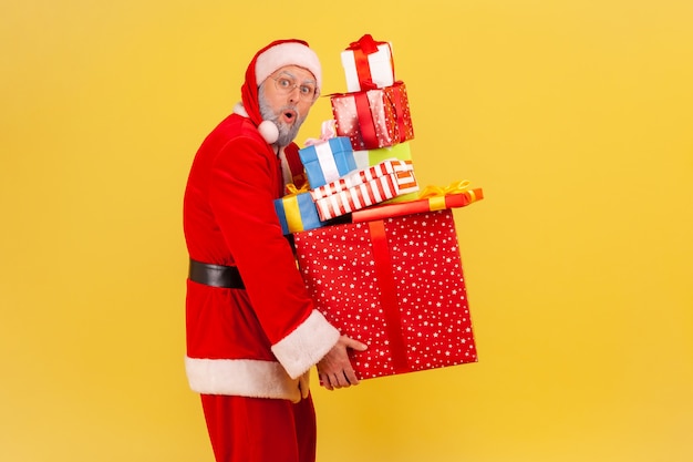 Santa claus sosteniendo una pesada pila de regalos, mirando a la cámara con la boca abierta, vacaciones de invierno.