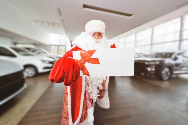 Foto santa claus sostiene una hoja en blanco con un lugar para el texto en el fondo de un concesionario de automóviles