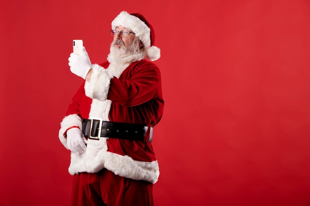 Santa Claus tomando un selfie con un teléfono móvil sobre fondo rojo.