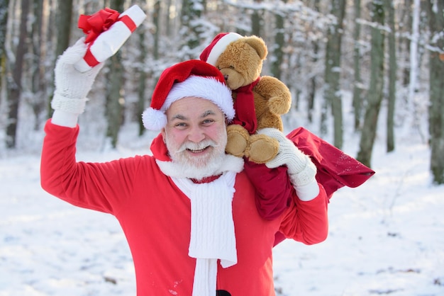 Santa divertido con regalo y oso de peluche en la víspera de Navidad fuera