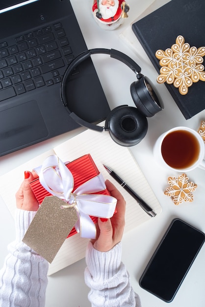 Santa secreto. Celebración de Navidad en la oficina. Escritorio de oficina con ordenador portátil, blocs de notas, material de oficina, smartphone y caja de regalo de Navidad grande con lazo festivo. flatlay