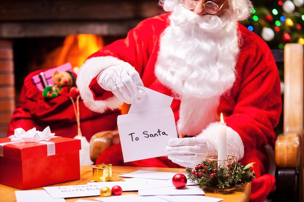 Santa en el trabajo. Close-up de Santa Claus leyendo cartas mientras está sentado en su silla con el árbol de Navidad una chimenea en el fondo