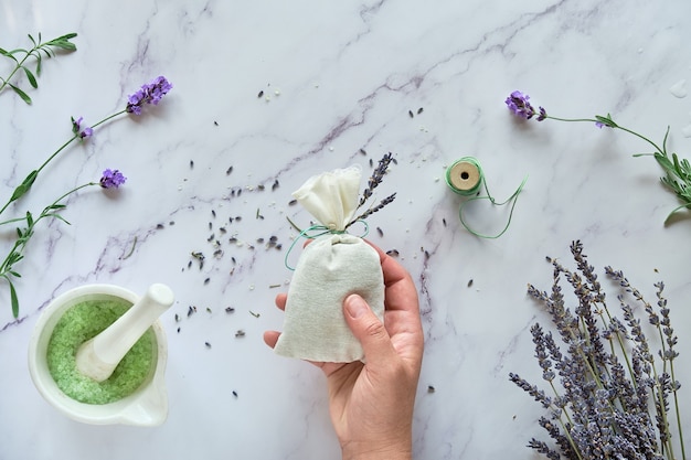 Saquinhos artesanais de lavanda e esfoliante de açúcar caseiro. Mão segura um saco de linho com flores secas de lavanda. Plano deitado em mármore branco.