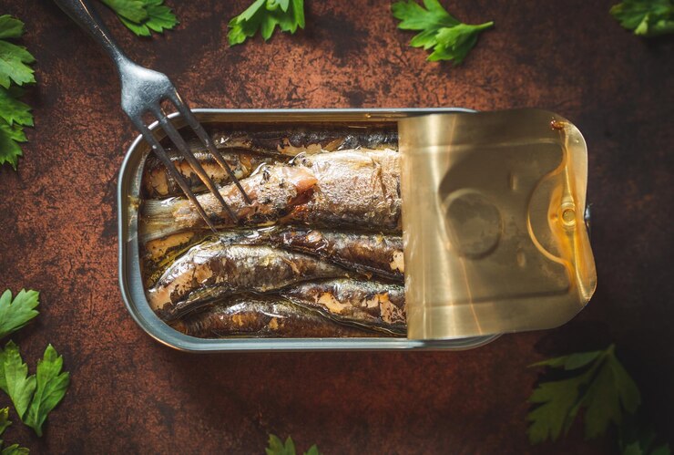 Foto sardinha enlatada com azeite aberta sobre uma mesa marrom escura com salsa.