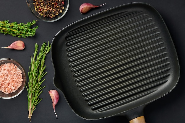 Sartén cuadrada vacía sobre fondo de mesa negro con especias y condimentos. Utensilios de plancha para asar carne o verduras. Vista superior con espacio para copiar texto.