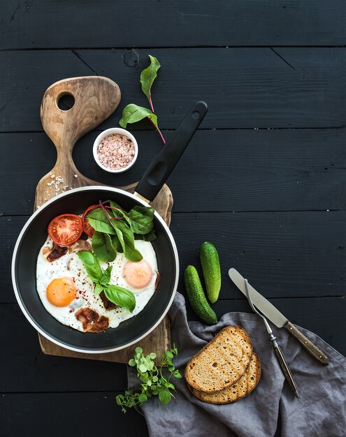 Sartén de huevos fritos, tocino, tomates con pan, remolacha forrajera y pepinos sobre tabla de madera rústica sobre la superficie de la mesa oscura