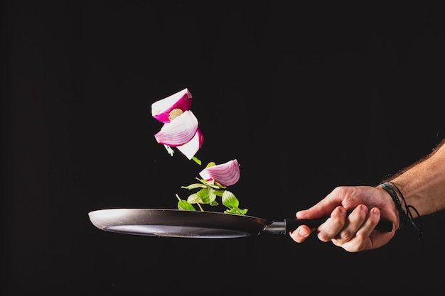 Sartén lanzando ingredientes como cebolla roja queso albahaca y condimentos en el aire