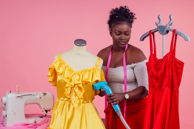 Sastre afroamericano modista con dispositivo de vapor generador de vapor caliente para vestido rojo y amarillo de seda en estudio de salón de tintorería fondo rosa