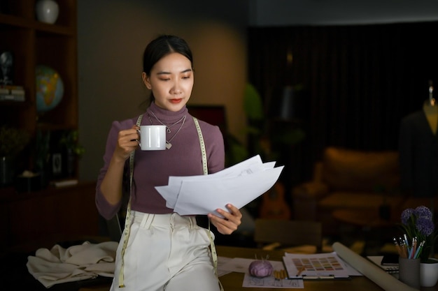 Una sastre asiática enfocada bebiendo café y revisando sus bocetos modelo en un estudio