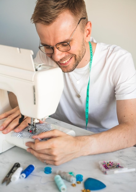 Un sastre con una cinta métrica en el cuello con una camiseta blanca Un hombre rubio sonriente con anteojos está cosiendo en un taller casero El concepto de estilo de vida de las pequeñas empresas y disfrutando del trabajo