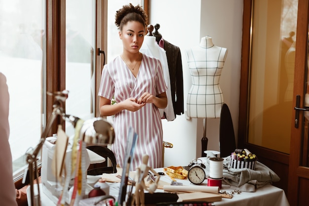 Sastre creativo trabajador. Joven africana, cerca de la mesa de trabajo con diferentes artículos de artesanía. Ella de pie cerca del maniquí de vestir