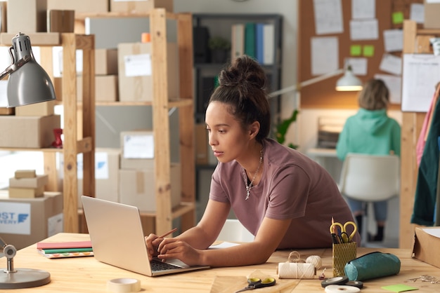 Sastre joven escribiendo en la computadora portátil en la mesa durante su trabajo en el taller