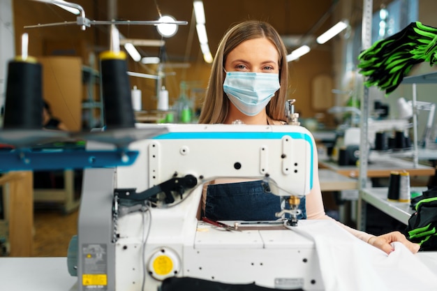 Sastre de mujer caucásica joven que trabaja en la fábrica de costura con máscara médica protectora.