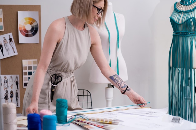 Sastre sonriente mide hilo verde con cinta adhesiva sobre la mesa en el trabajo de diseñadora de estudio de moda