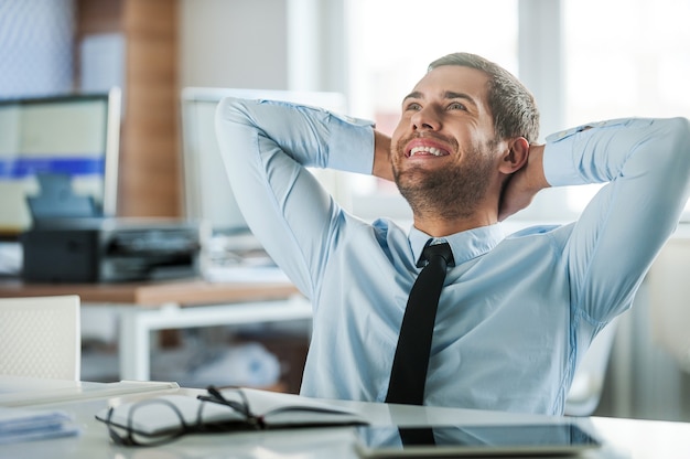 Foto satisfacción laboral. hombre de negocios alegre en ropa formal sosteniendo la cabeza entre las manos mientras está sentado en su escritorio en la oficina